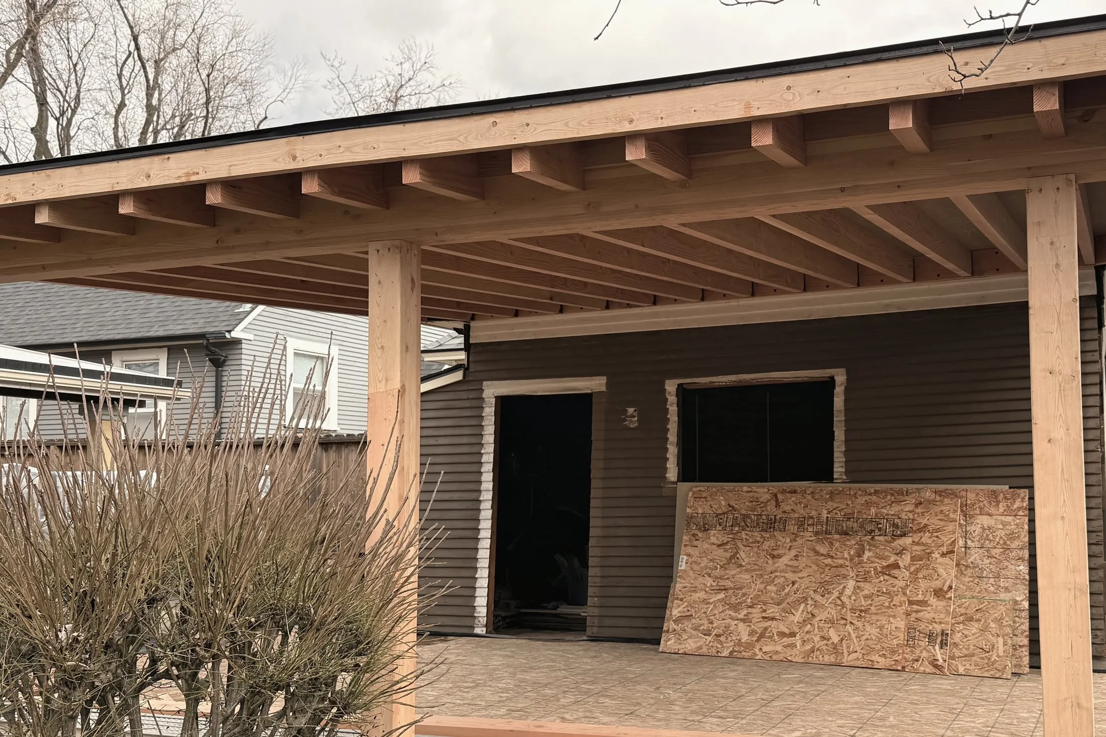 The carport standing — posts, beams, and rafter tails, the roof running off the existing garage